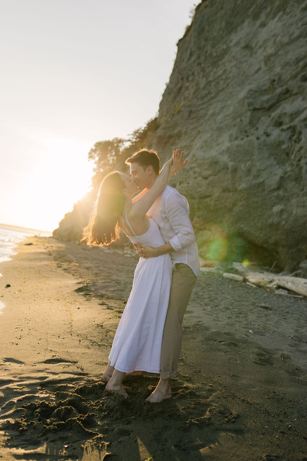 A romantic couple embraces on a beach at sunset with dramatic cliffs and golden light creating a dreamy atmosphere.