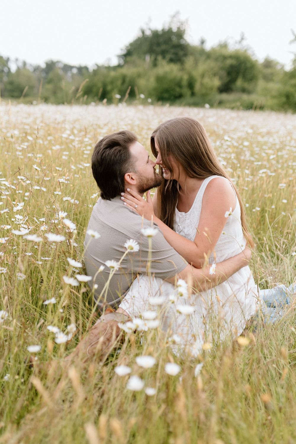 A romantic couple in white outfits share an intimate moment while sitting in a dreamy meadow of wildflowers.