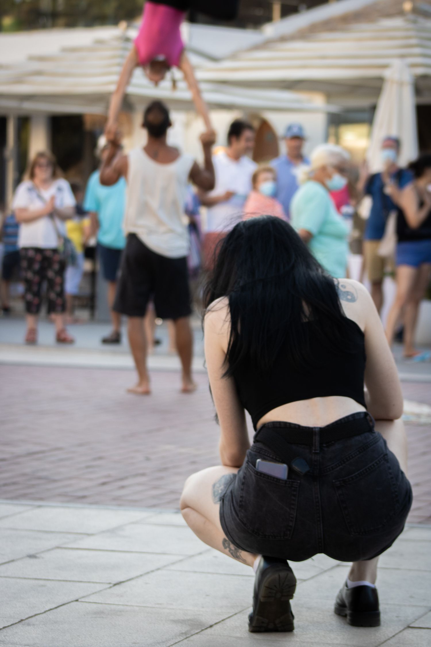 Person crouching low to take a photo on a paved outdoor surface while others walk in the background.