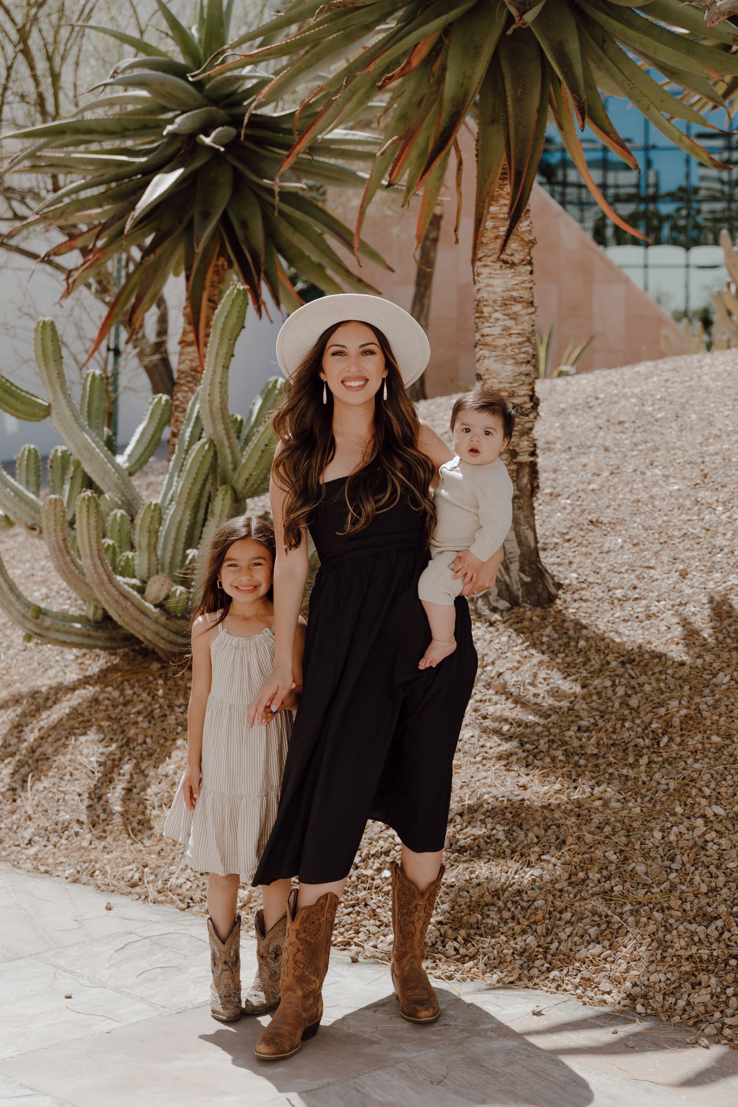 Woman in black dress and cowboy boots with two children posing by desert plants.
