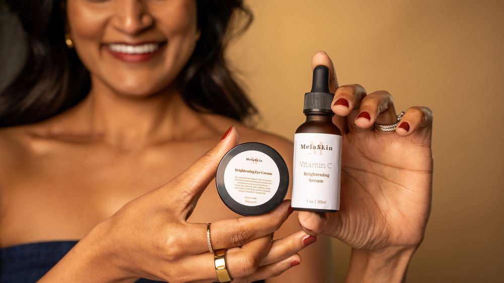 Someone holds up skincare products including a dark jar and dropper bottle against a warm brown background.
