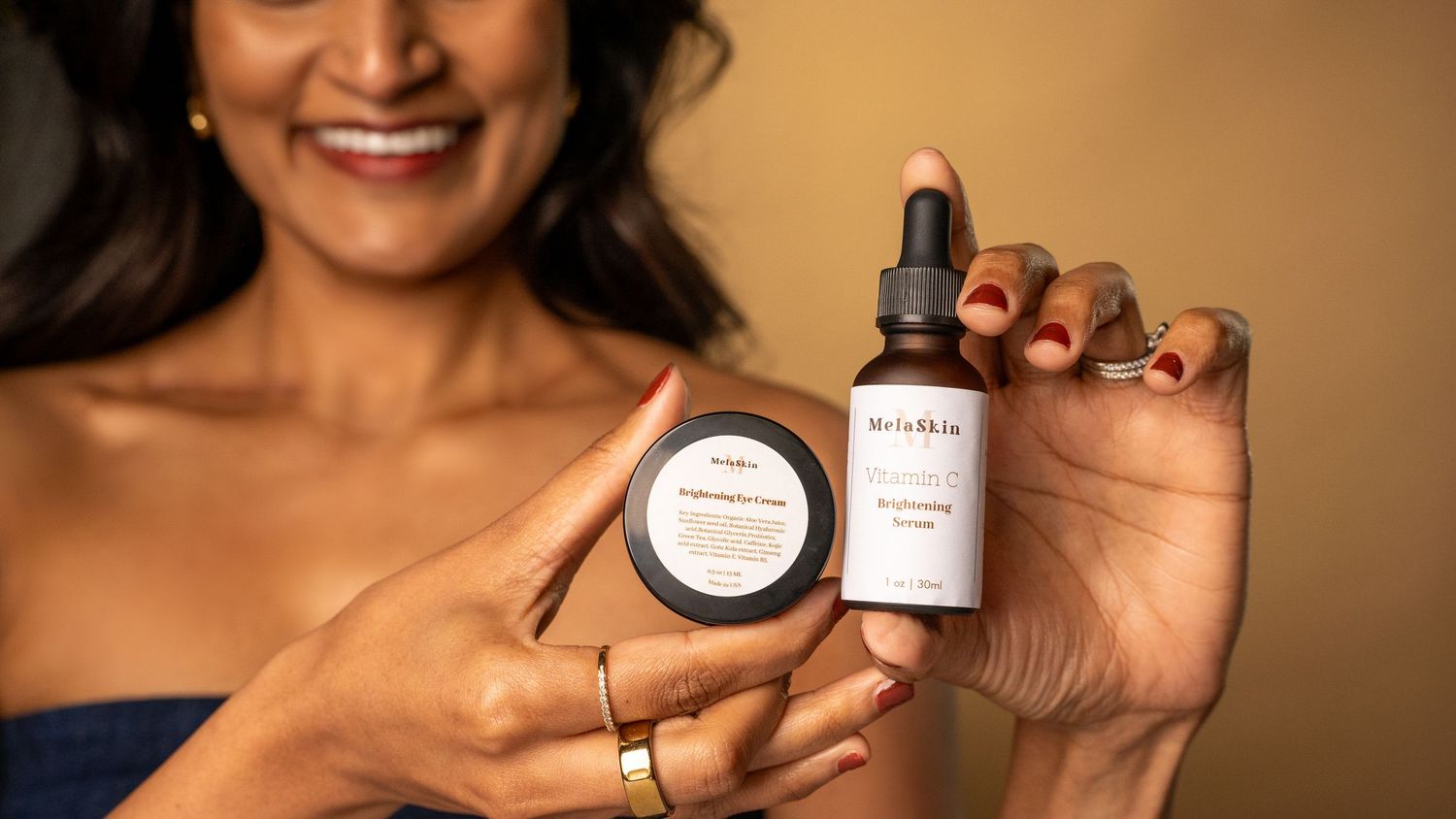 Someone holds up skincare products including a dark jar and dropper bottle against a warm brown background.