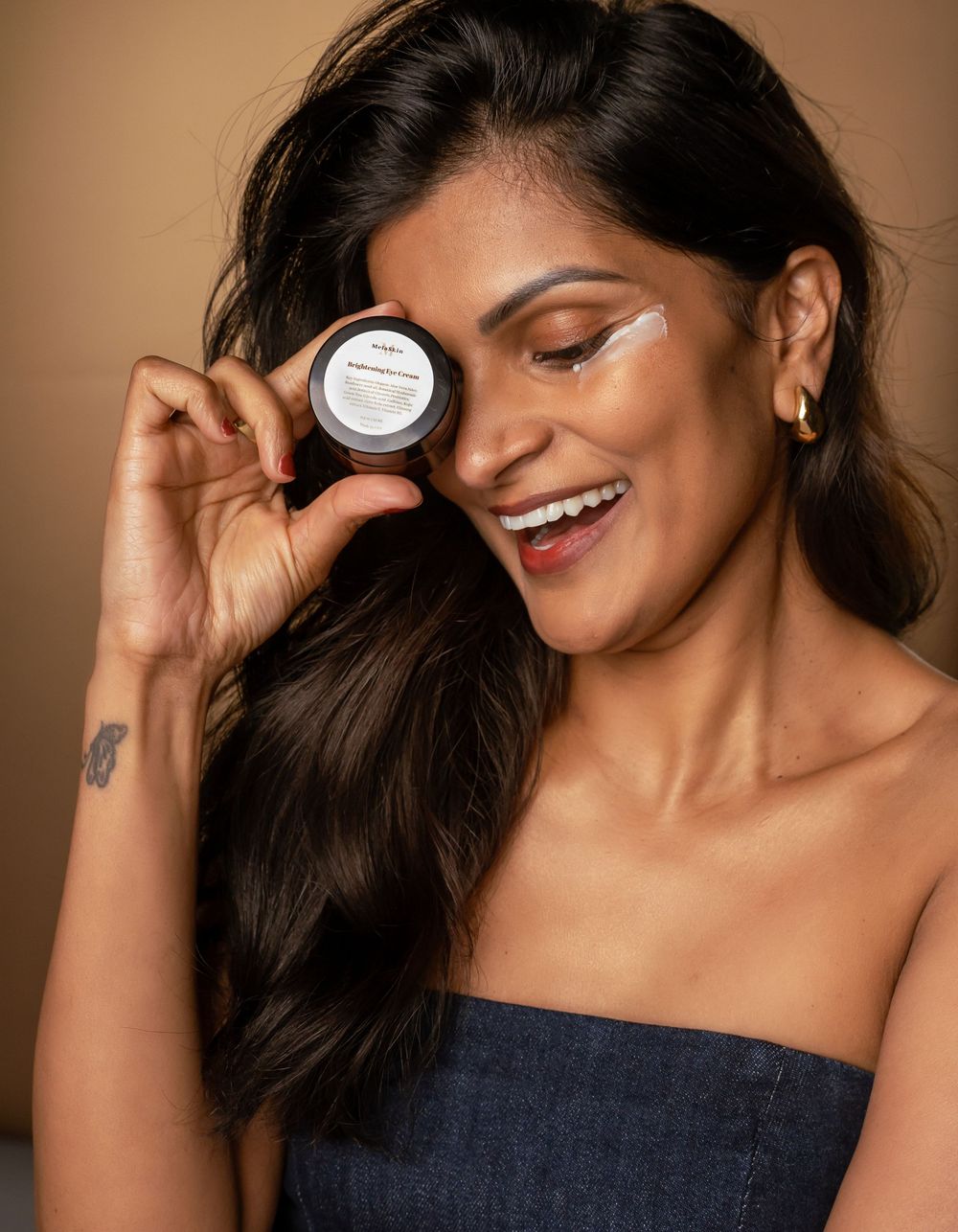 Smiling model in denim strapless top holds up a beauty product container while posing against brown backdrop.