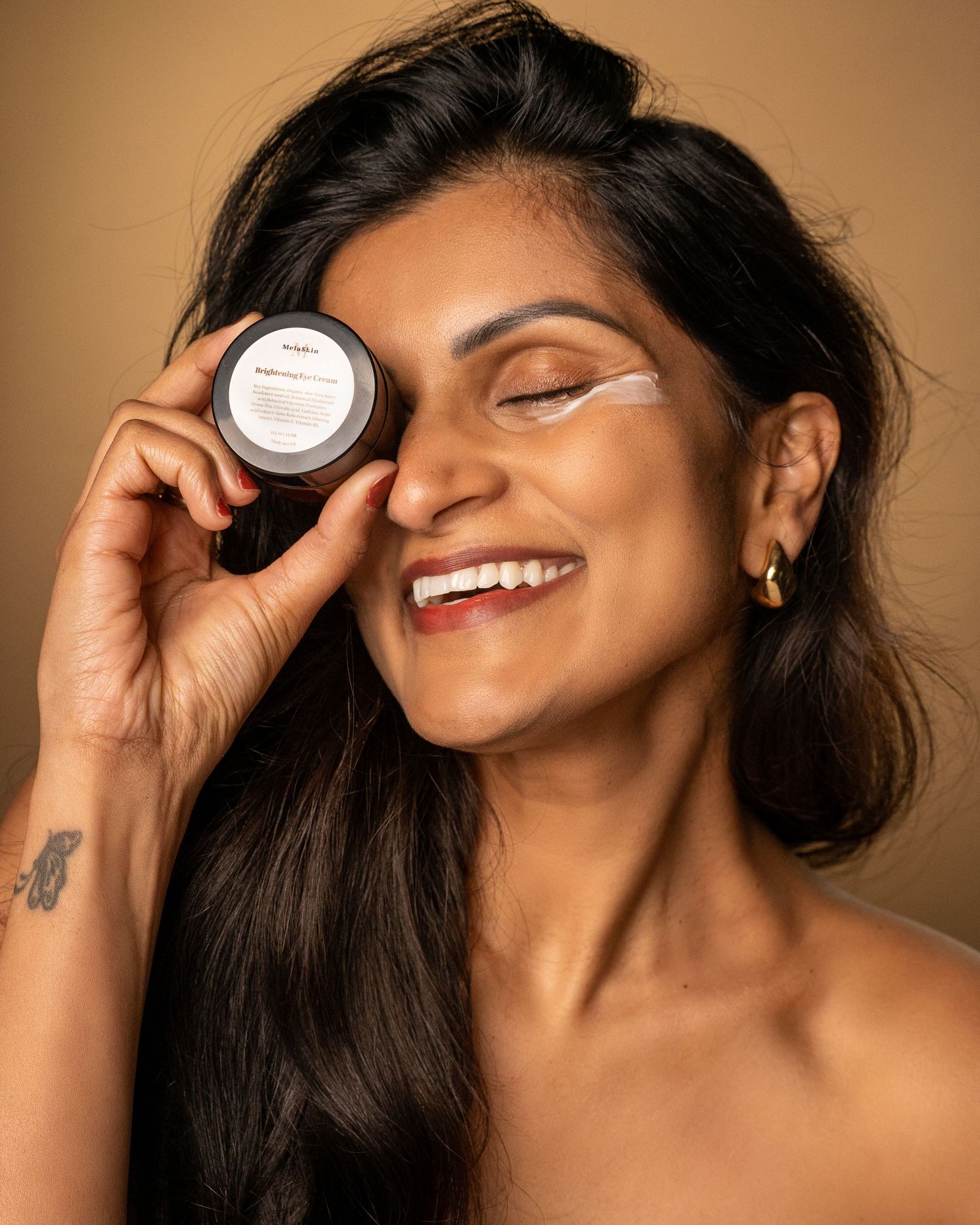 Smiling woman with long dark hair holds a beauty product container near her face against a warm brown background.