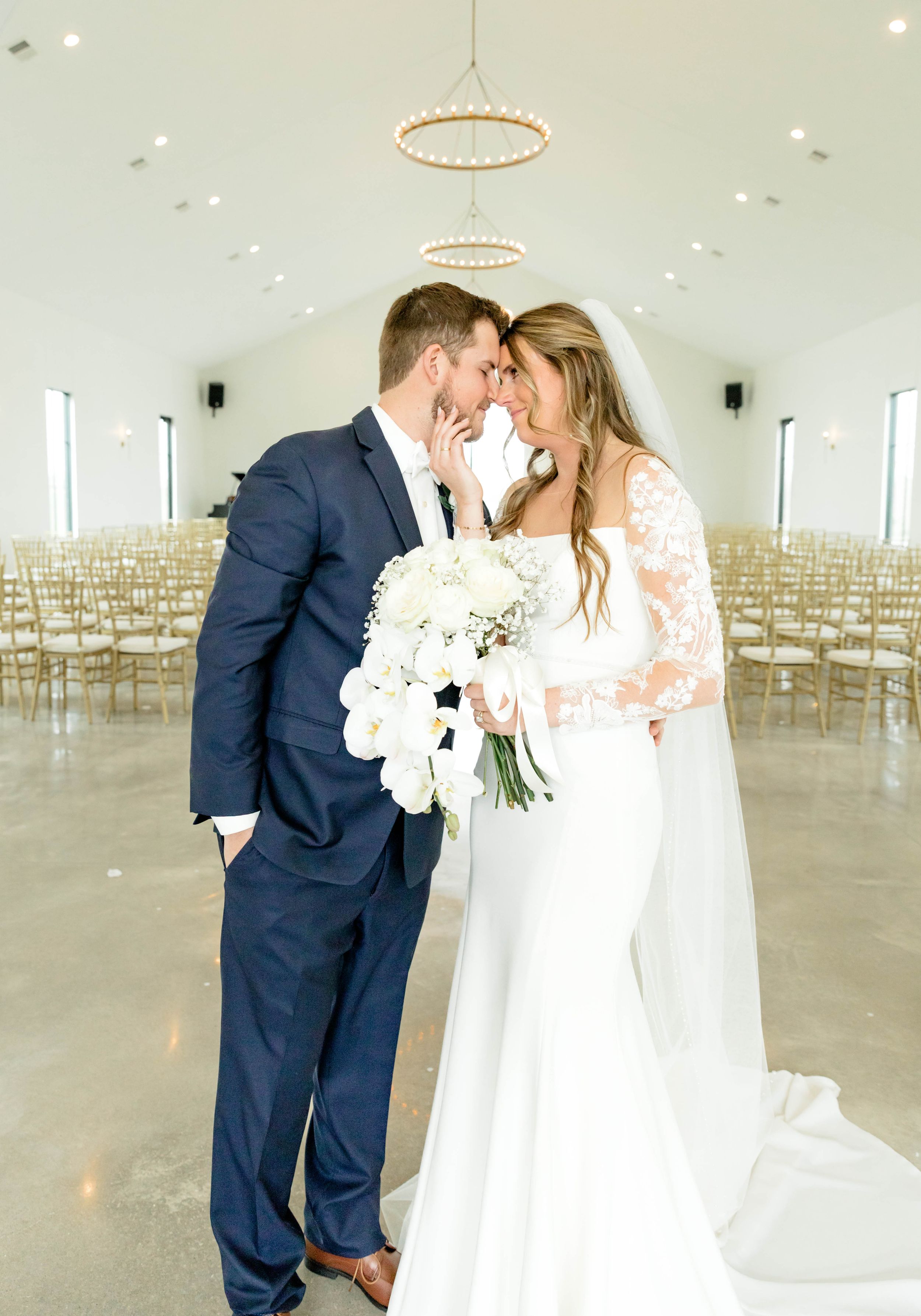 A romantic wedding moment captured in a bright white industrial venue with elegant circular chandeliers and gold chairs.