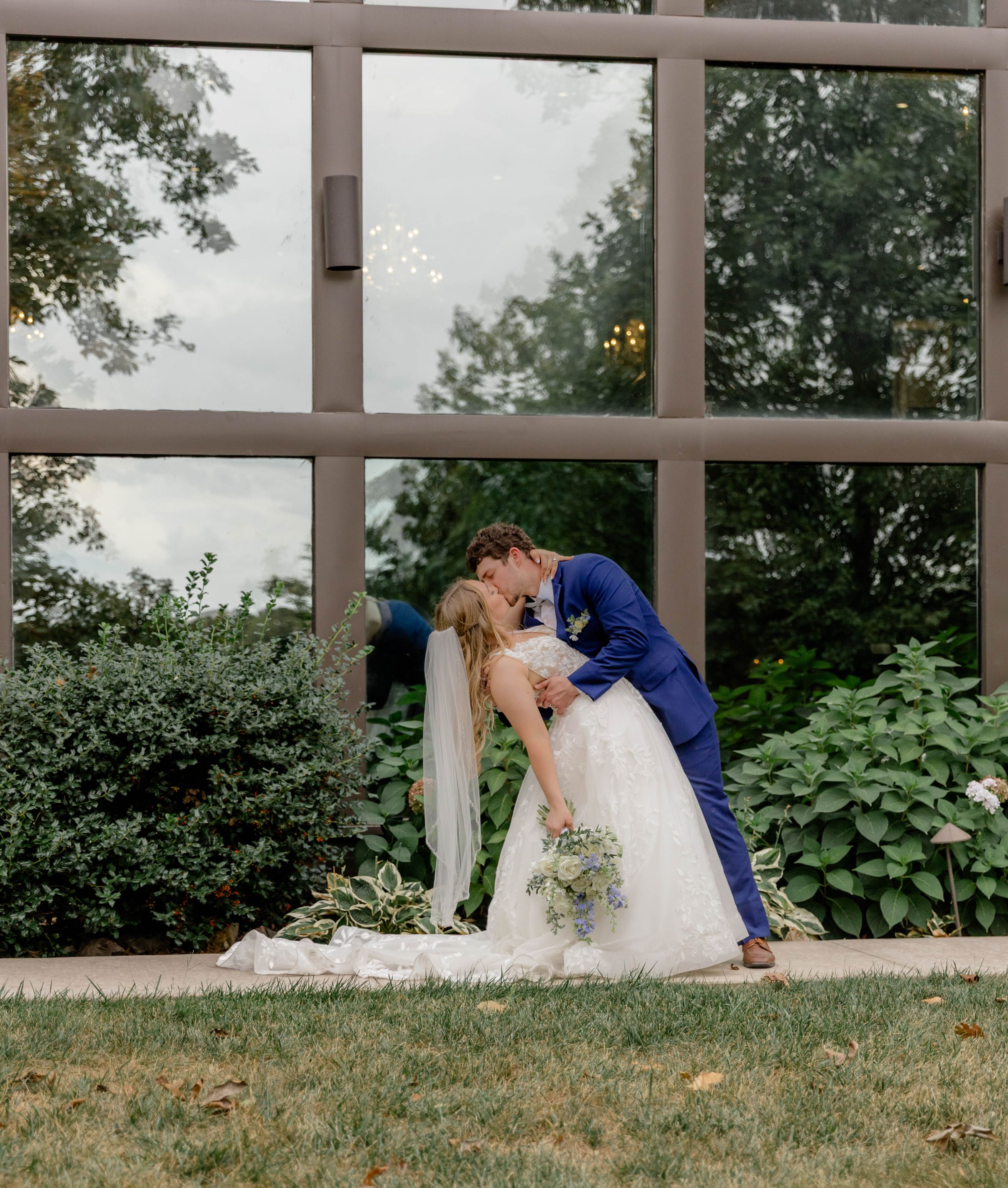 A couple shares a romantic dip kiss in front of a large window overlooking lush greenery on their wedding day.