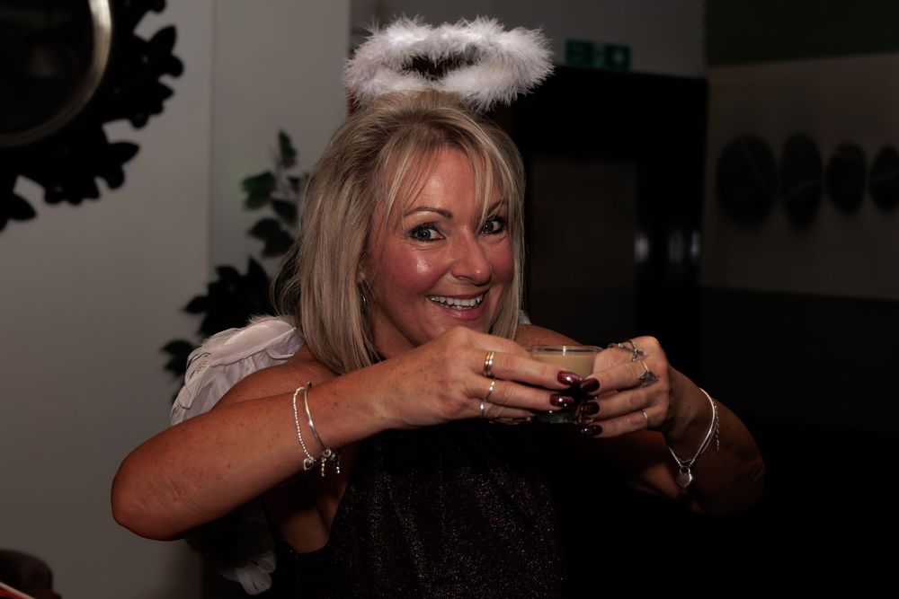 Woman in angel costume with halo headband smiling and holding a drink at a Halloween party.
