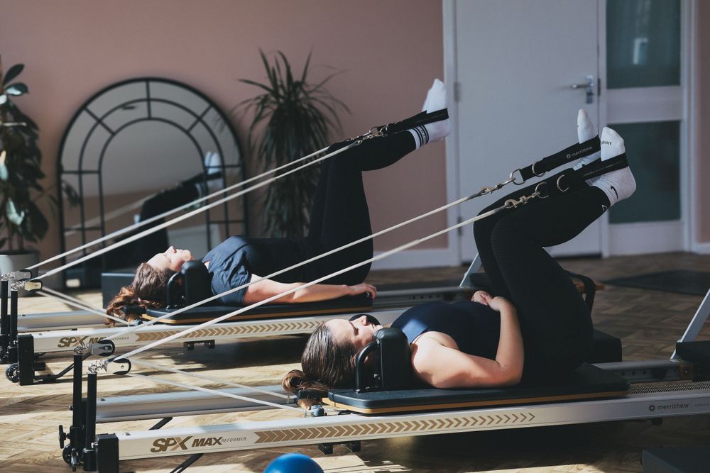 Two women lying on SPX Max reformer machines doing Pilates leg exercises.