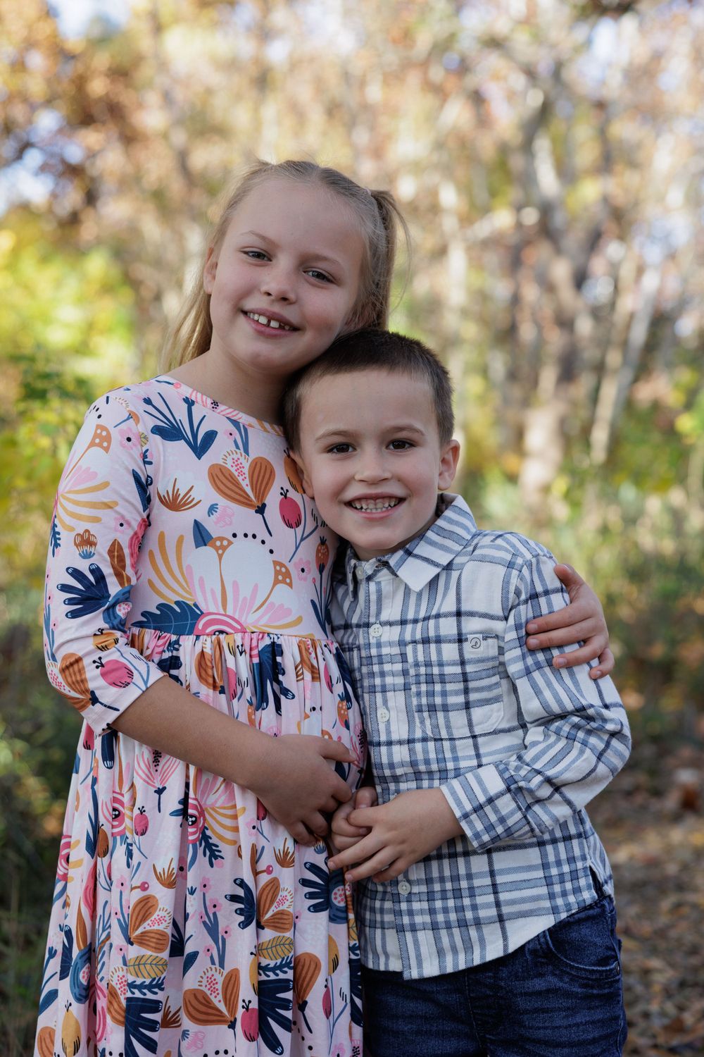 Two children stand close together smiling in a wooded outdoor setting.