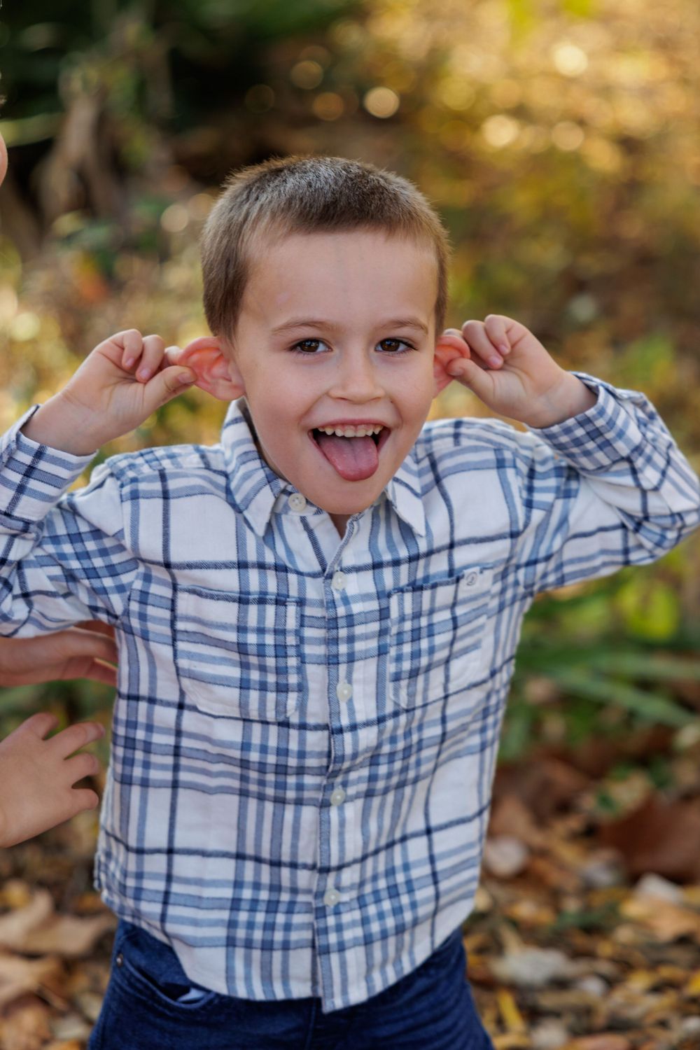 A young child in a plaid shirt makes a silly face while pulling on their ears.