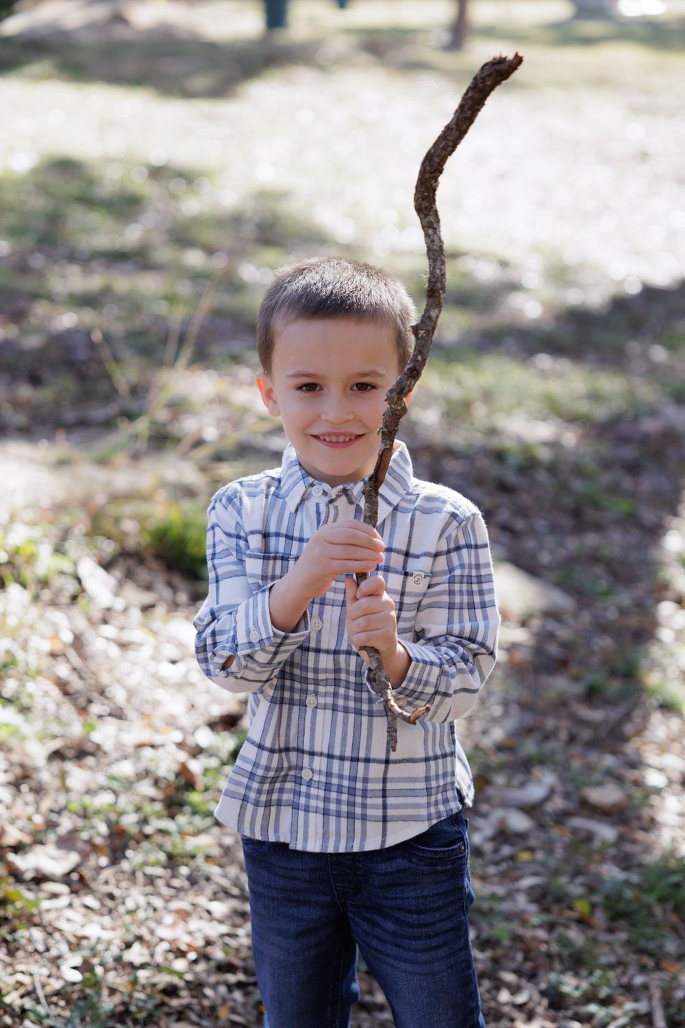 A young child in a plaid shirt holds up a large stick while standing in dappled sunlight.