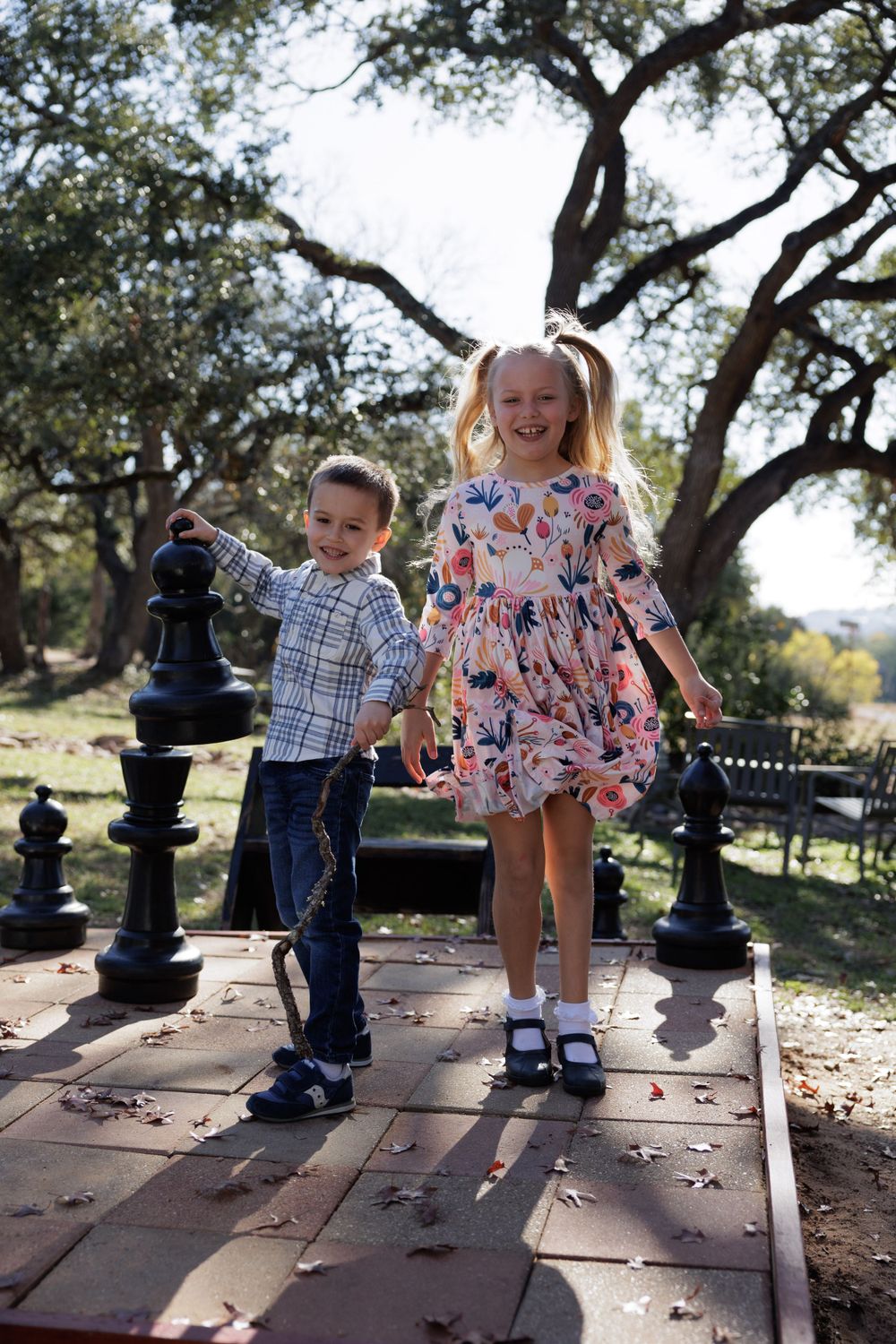 Two children play together on stone pavers under oak trees with large chess pieces in a scenic outdoor setting.