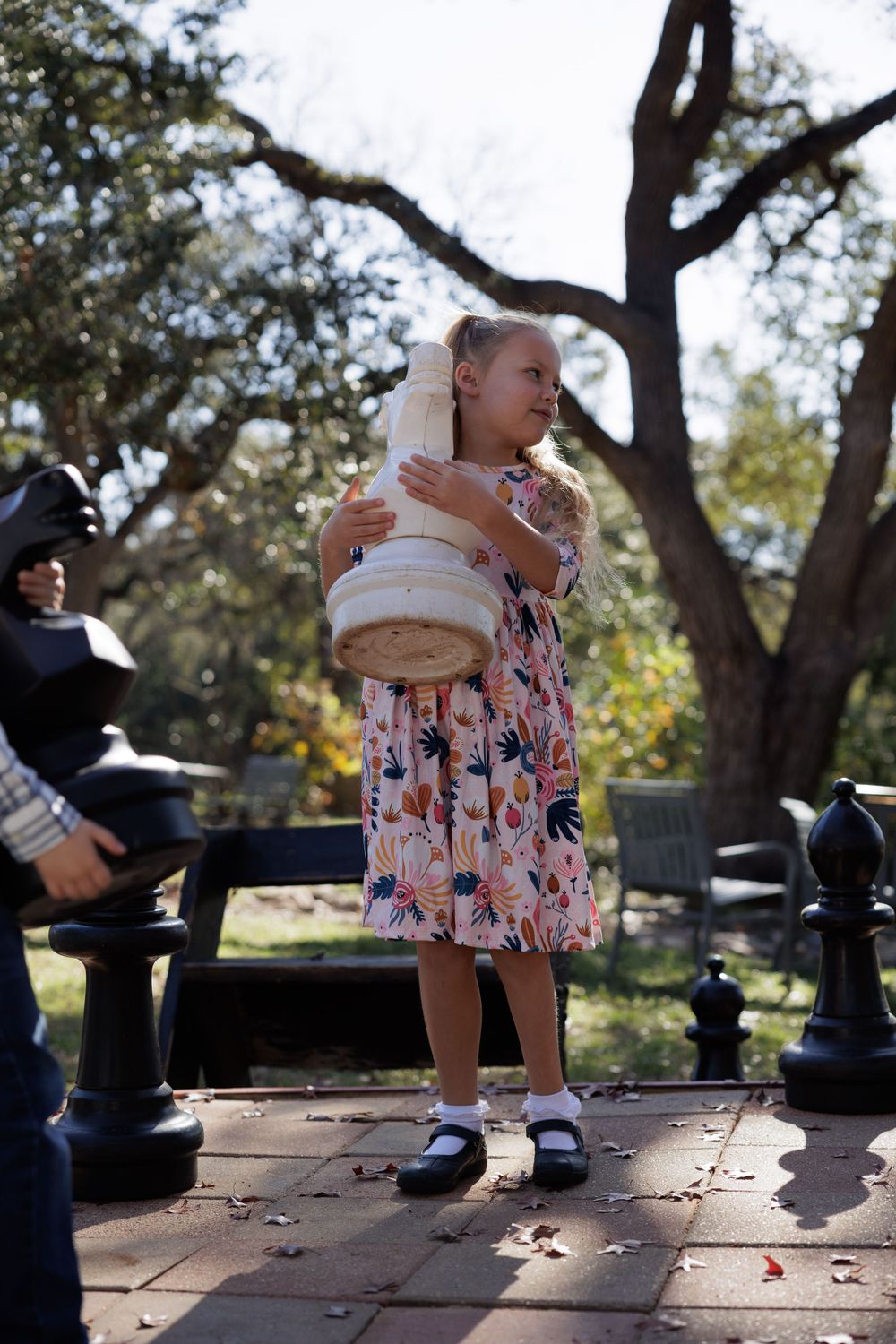 Child in floral dress holding a plush toy standing next to decorative chess pieces in a park setting.