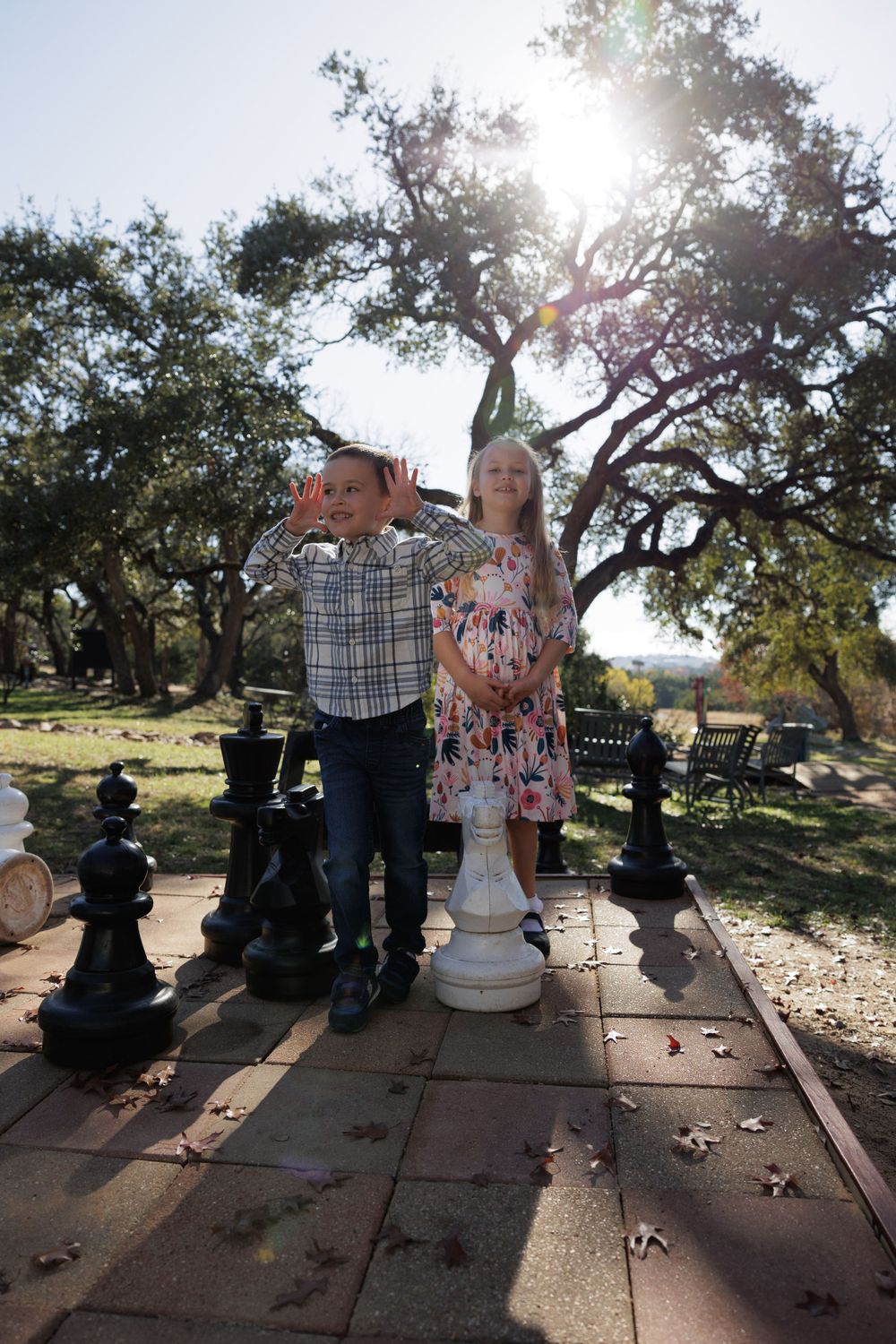 Backlit photo of children standing on stone pavers with large chess pieces under a majestic oak tree.