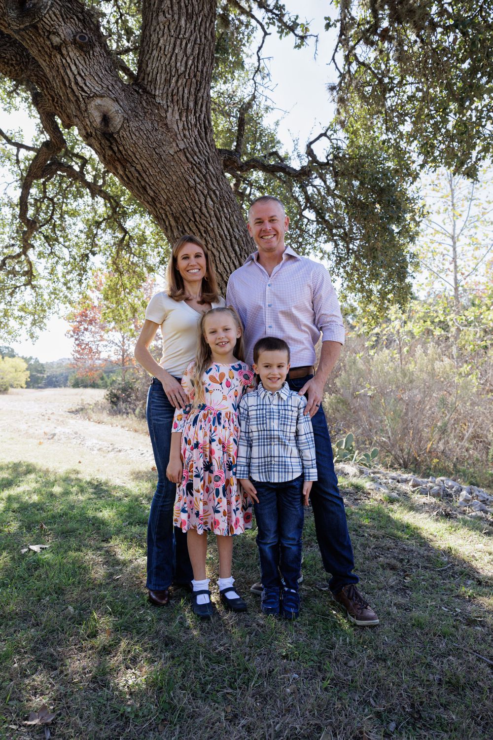 Family of four poses together under a large oak tree in casual attire.