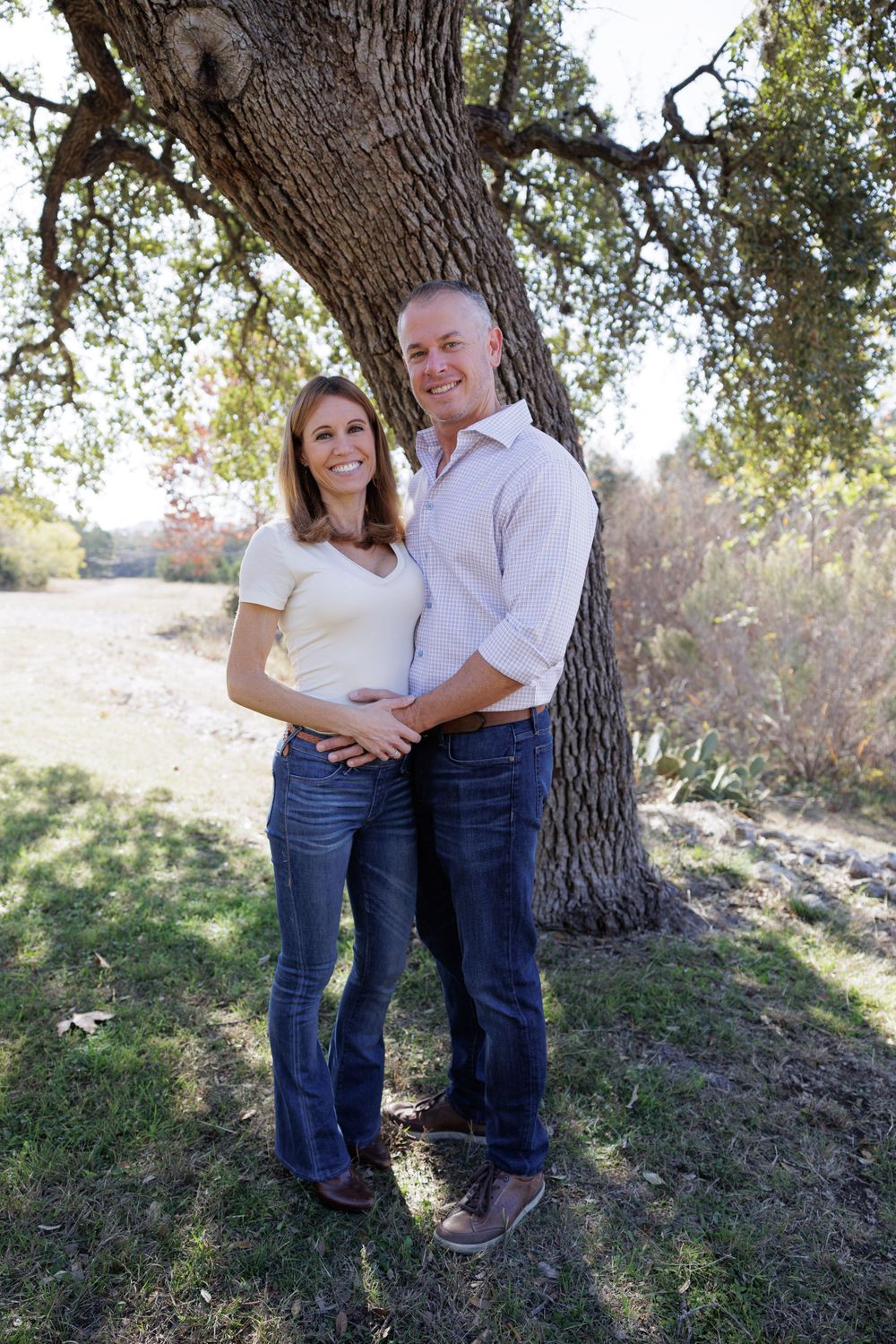 Couple in casual attire embracing while standing under a large oak tree in natural setting.