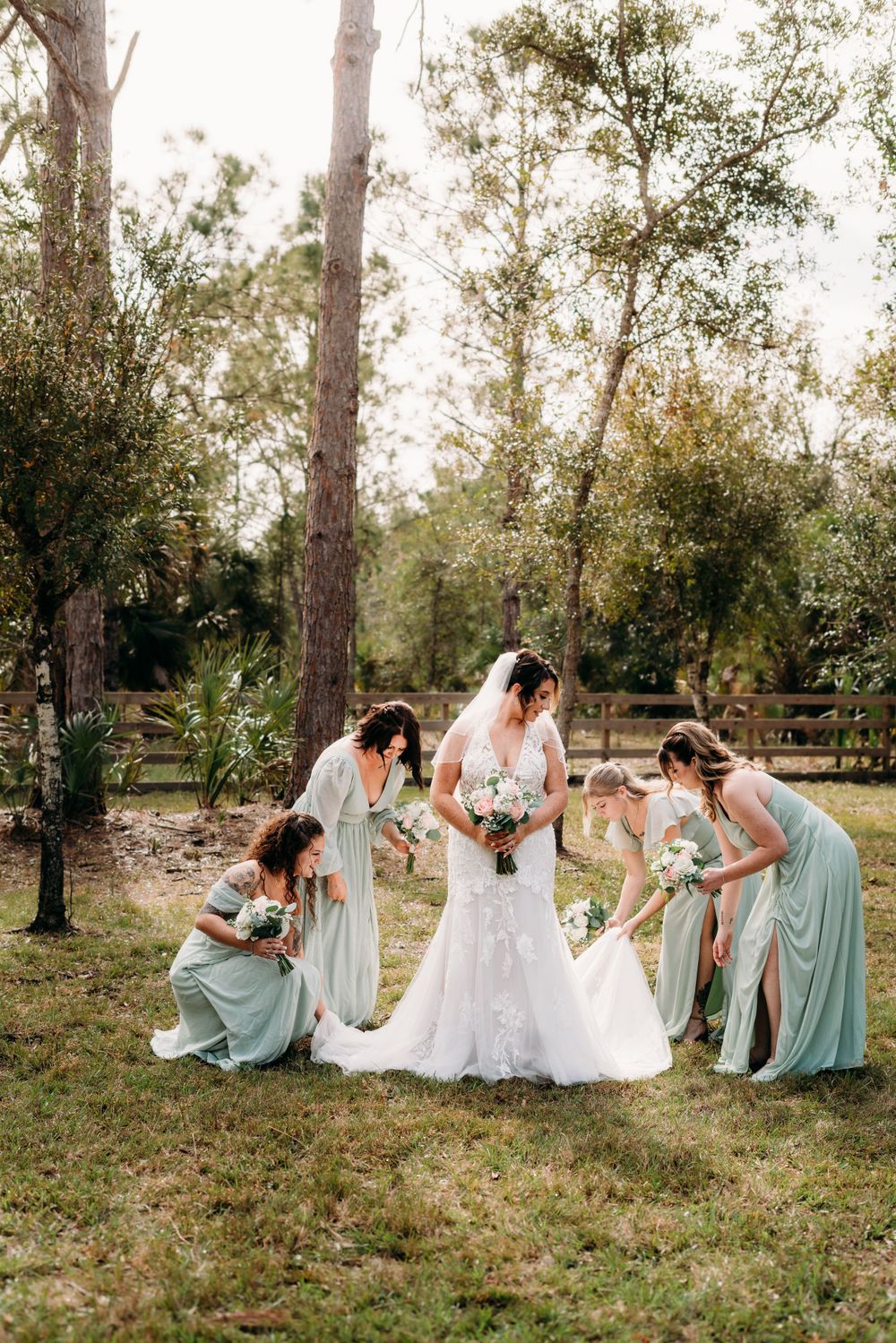 Bride looking down while bridesmaids fix her dress in a green field.
