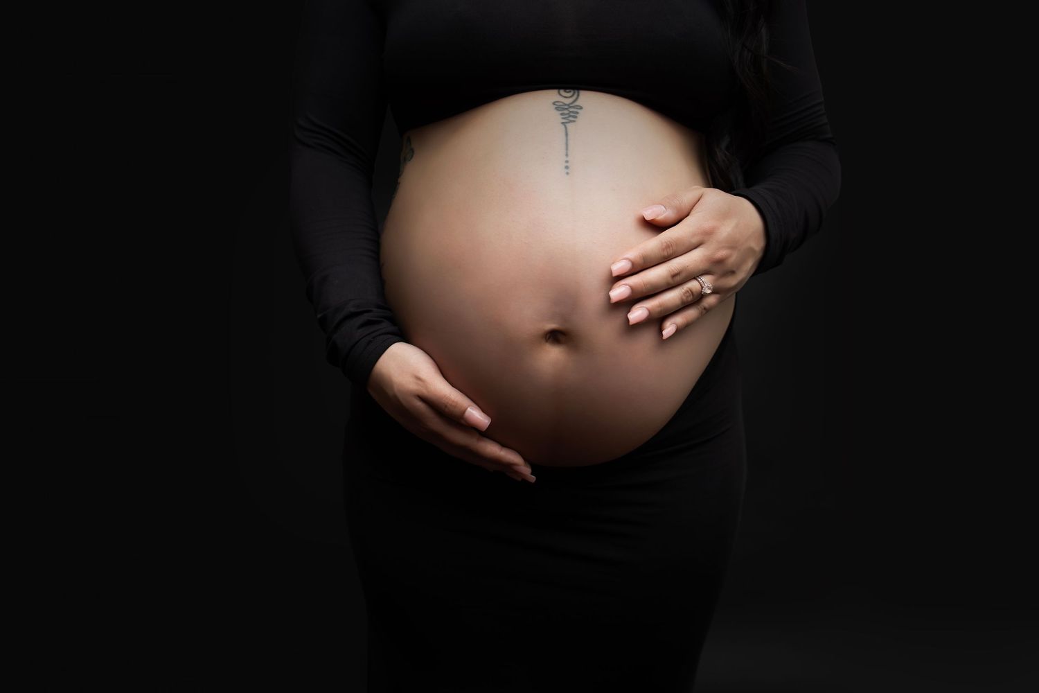 Close-up of pregnant belly with hands gently cradling bump against dark background.