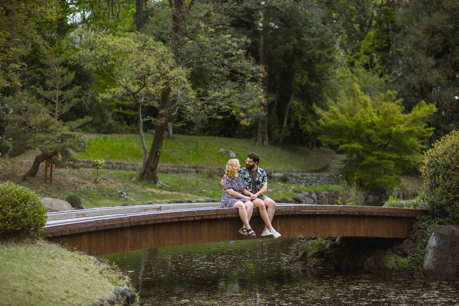 Couple sitting together on wooden bridge over pond in Japanese garden.