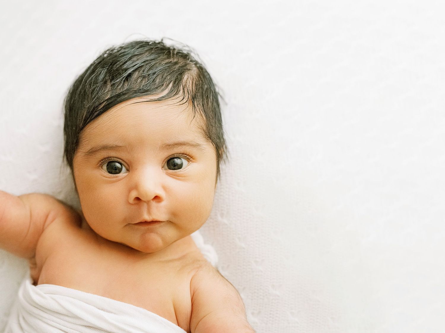 A small baby swaddled in white fabric looks at the camera with wide eyes against a light background.