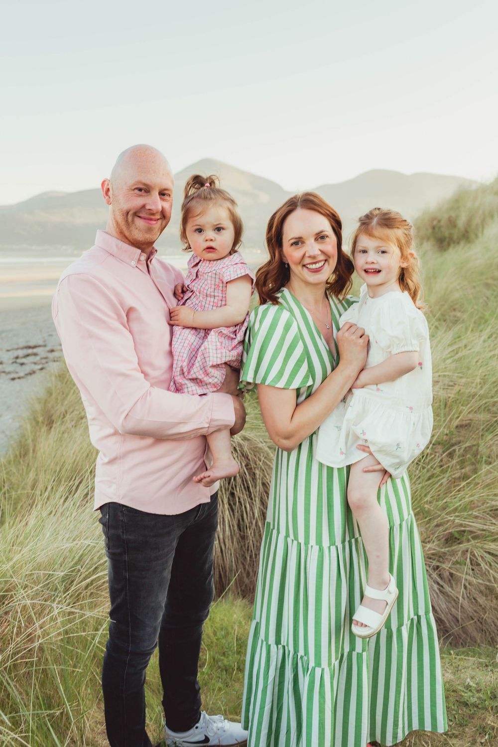 Family wearing coordinated pink and green outfits poses together on a beach with tall grass in the background.