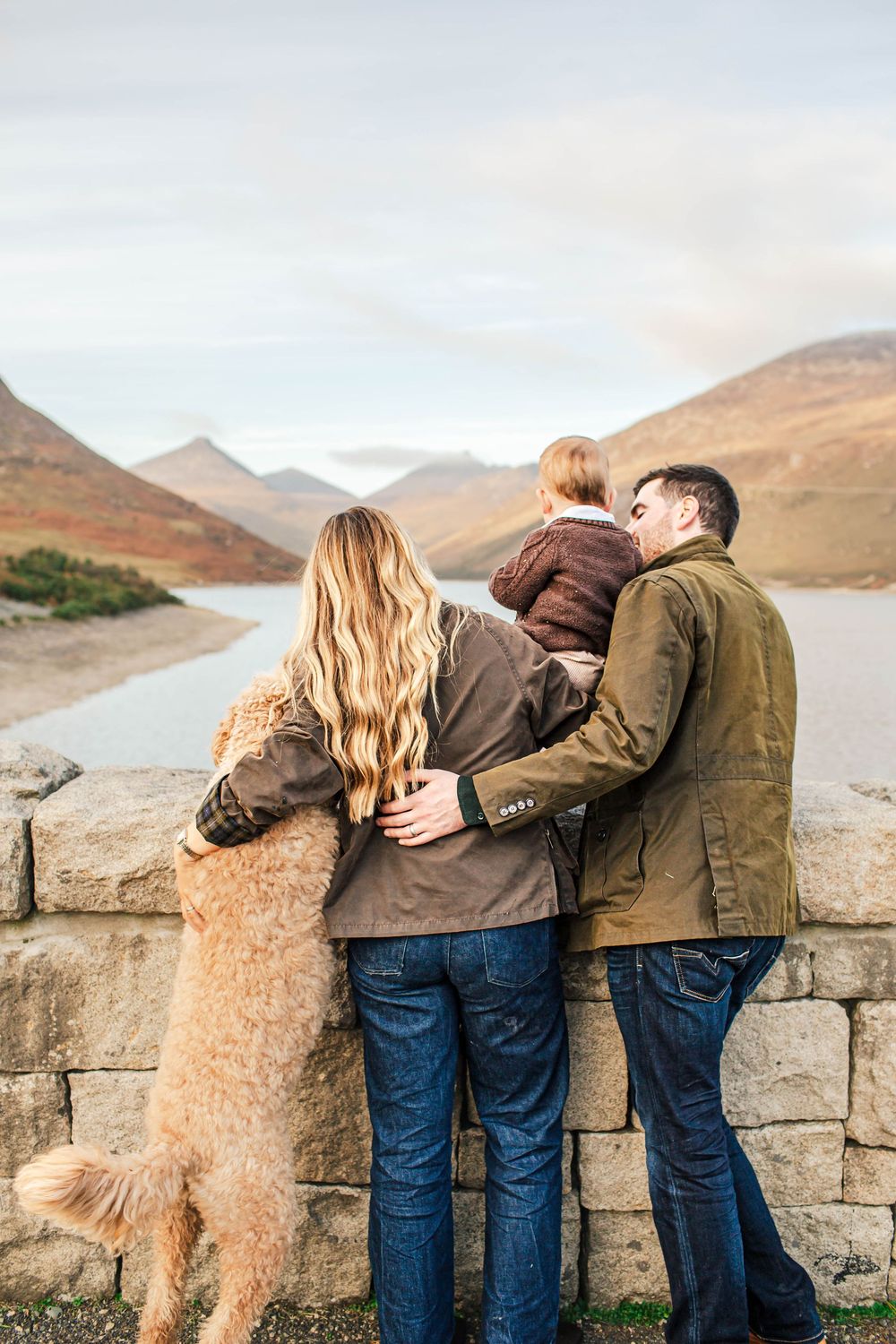 Family and golden retriever looking out over mountain lake while leaning on stone wall.