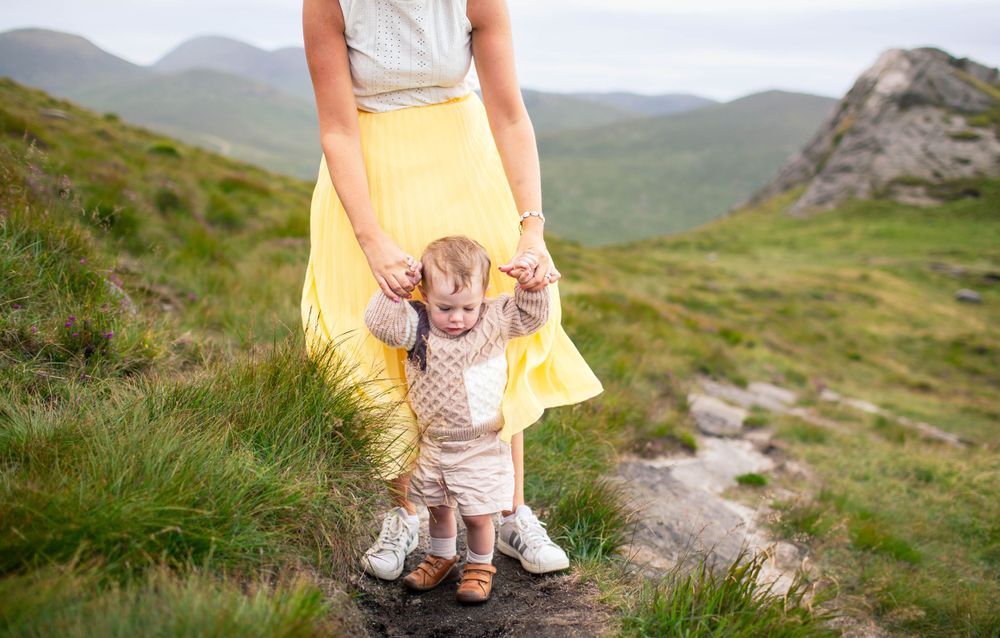 A yellow dress flows in the mountain breeze as a toddler stands supported on rocky terrain.