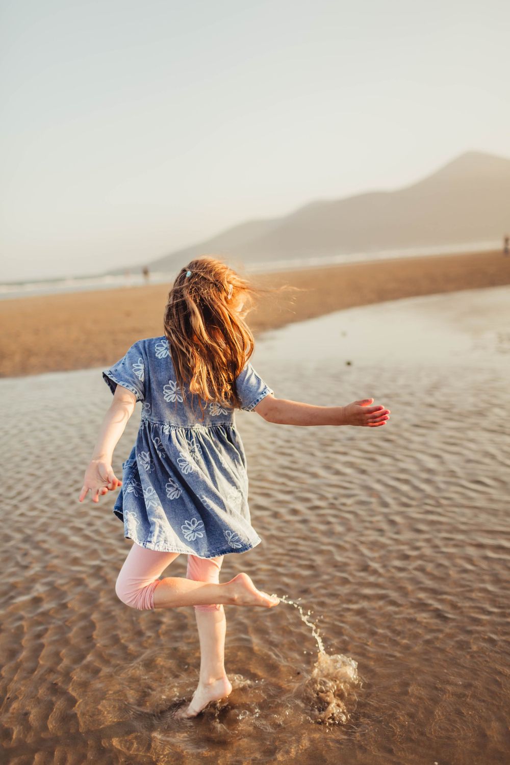 Young child in blue dress splashing and playing in shallow water at beach during golden sunset.