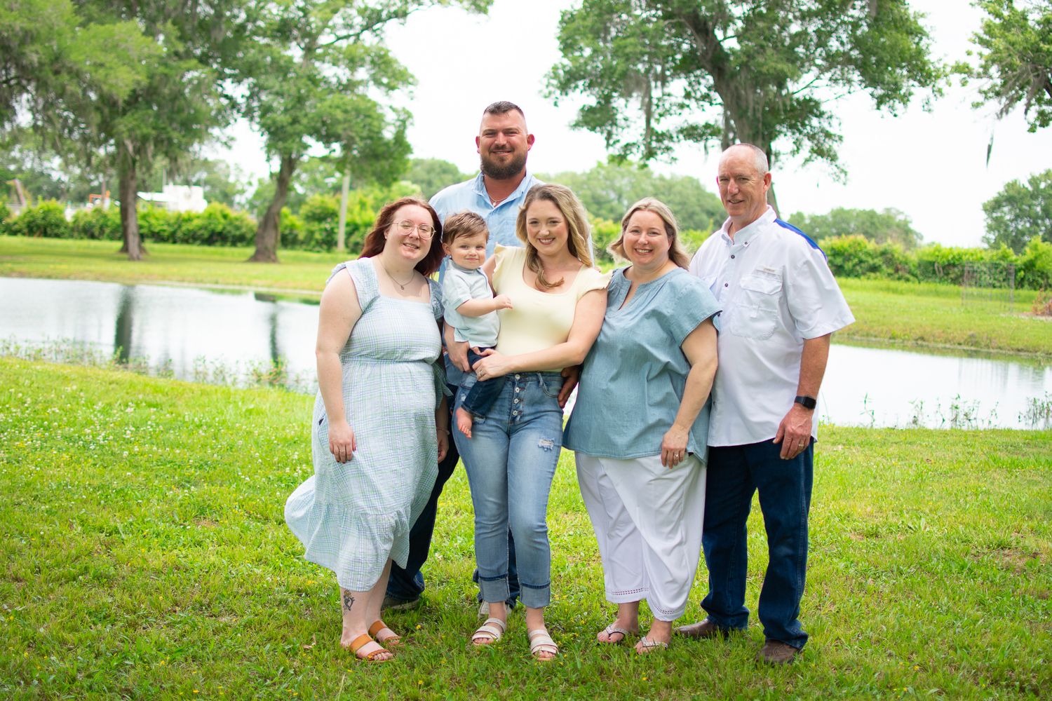 A cheerful family group poses together in casual attire by a pond on a sunny day.