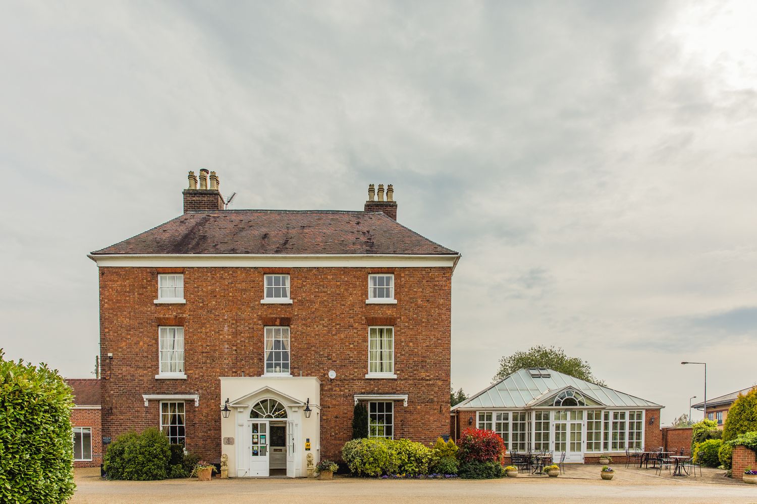 Elegant red brick Georgian manor house with white trim and conservatory extension under cloudy skies.