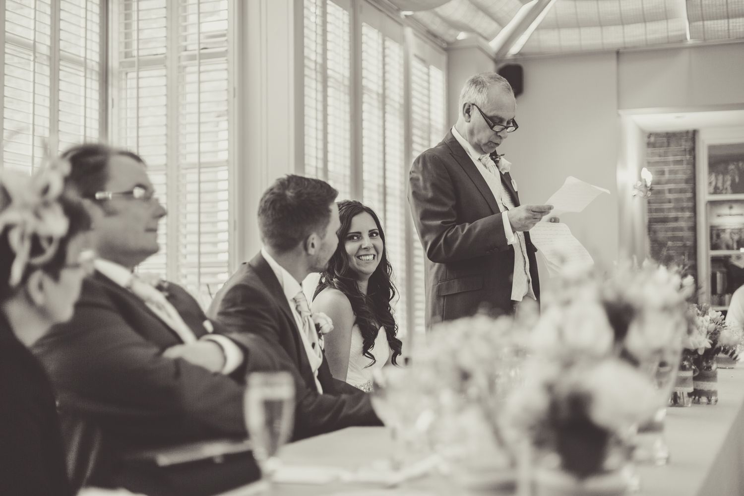 Black and white photo of wedding reception guests listening to a speech.
