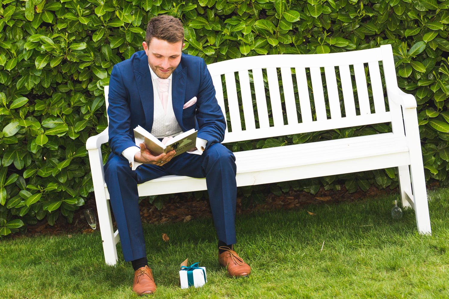 Man in navy suit reading book while sitting on white garden bench next to hedge.