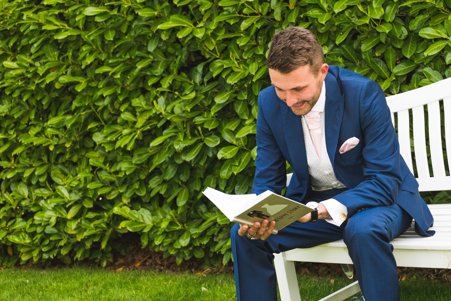 Groom in blue suit reading while relaxing on white outdoor bench against green hedge.