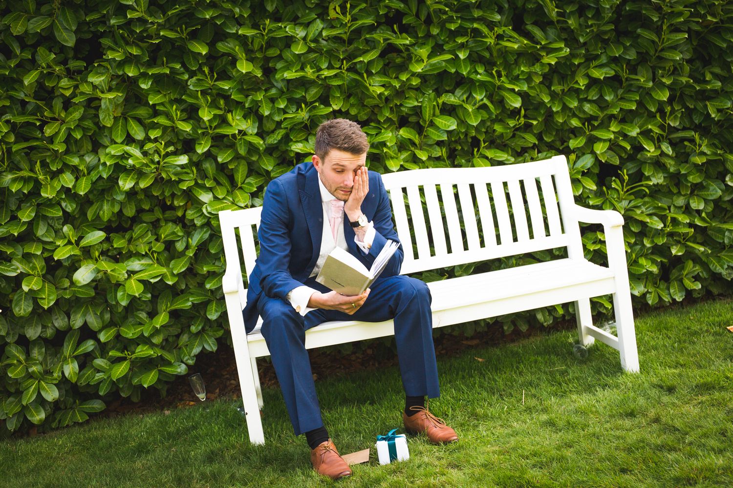 Man in blue suit sitting casually on white garden bench while reading.