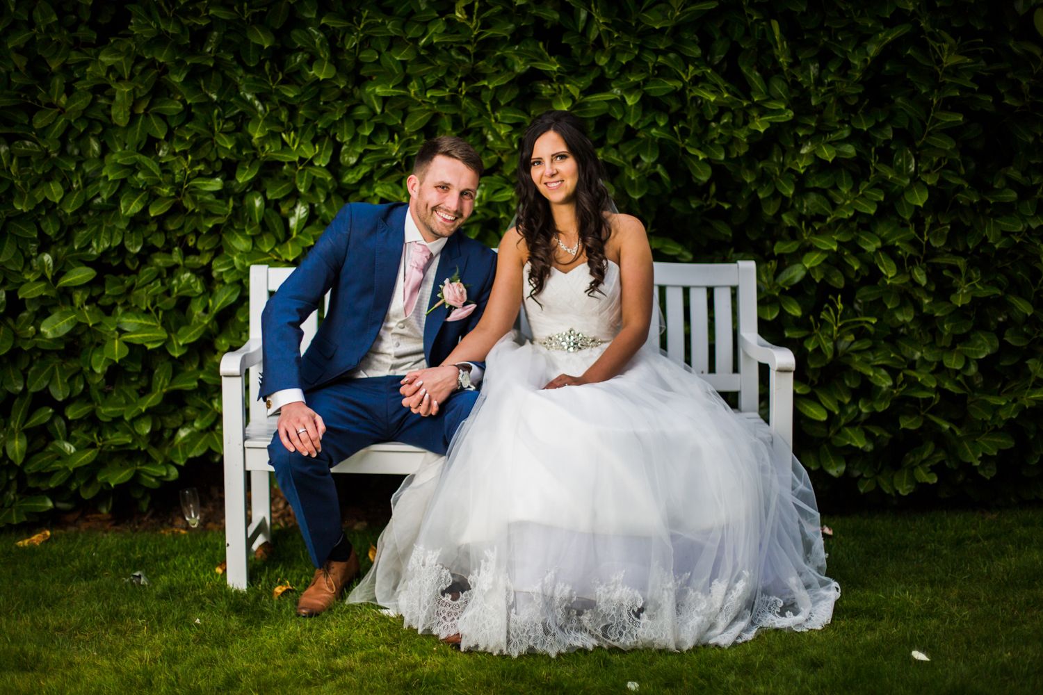 A wedding couple sits together on a white garden bench surrounded by lush green foliage.