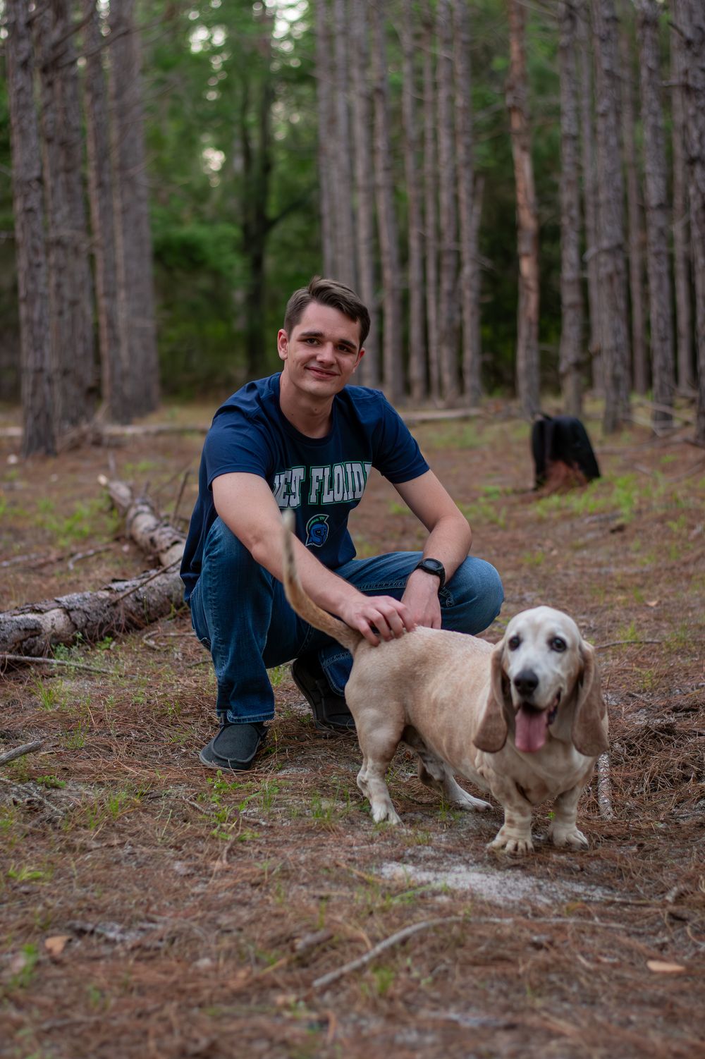 Man in blue t-shirt crouching with a golden Basset Hound in a pine forest setting.