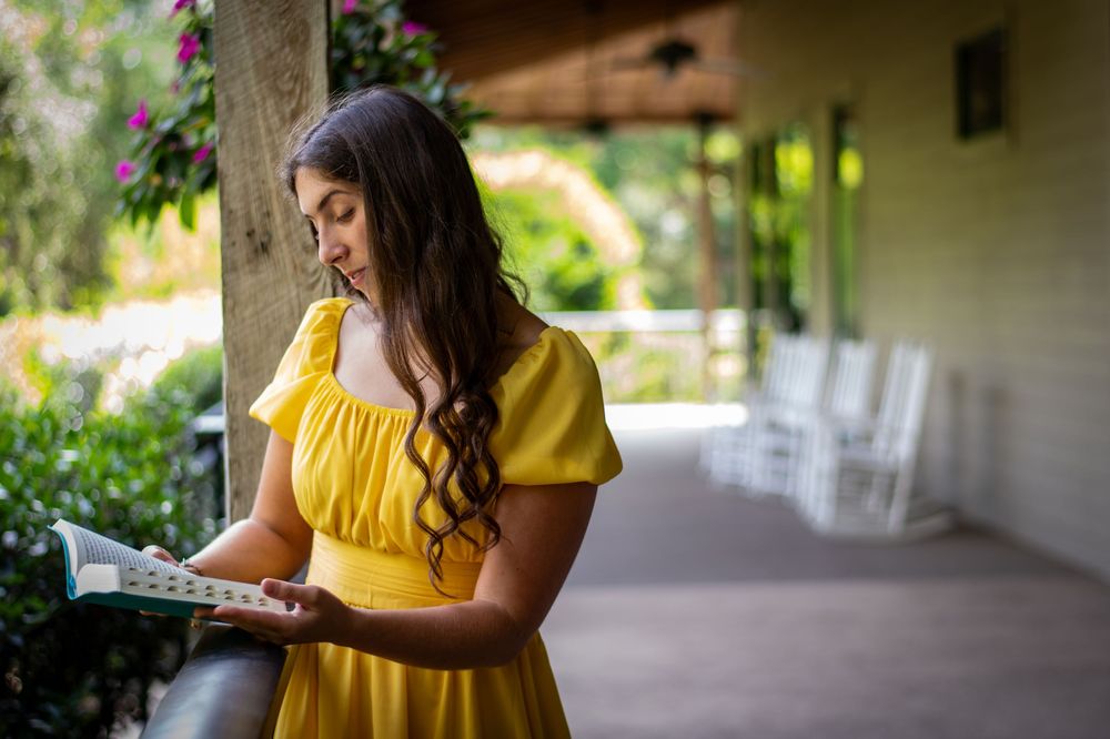 Young woman in yellow dress reading a book on a porch surrounded by lush greenery and pink flowers.