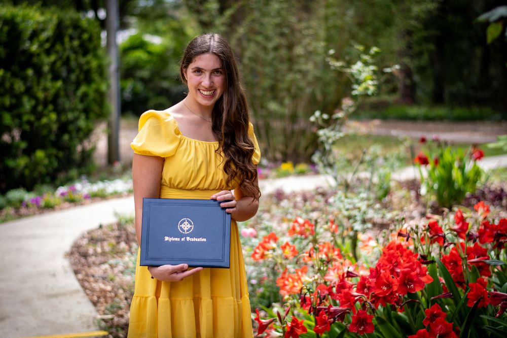 Young woman in yellow dress holding graduation diploma cover among colorful garden flowers.