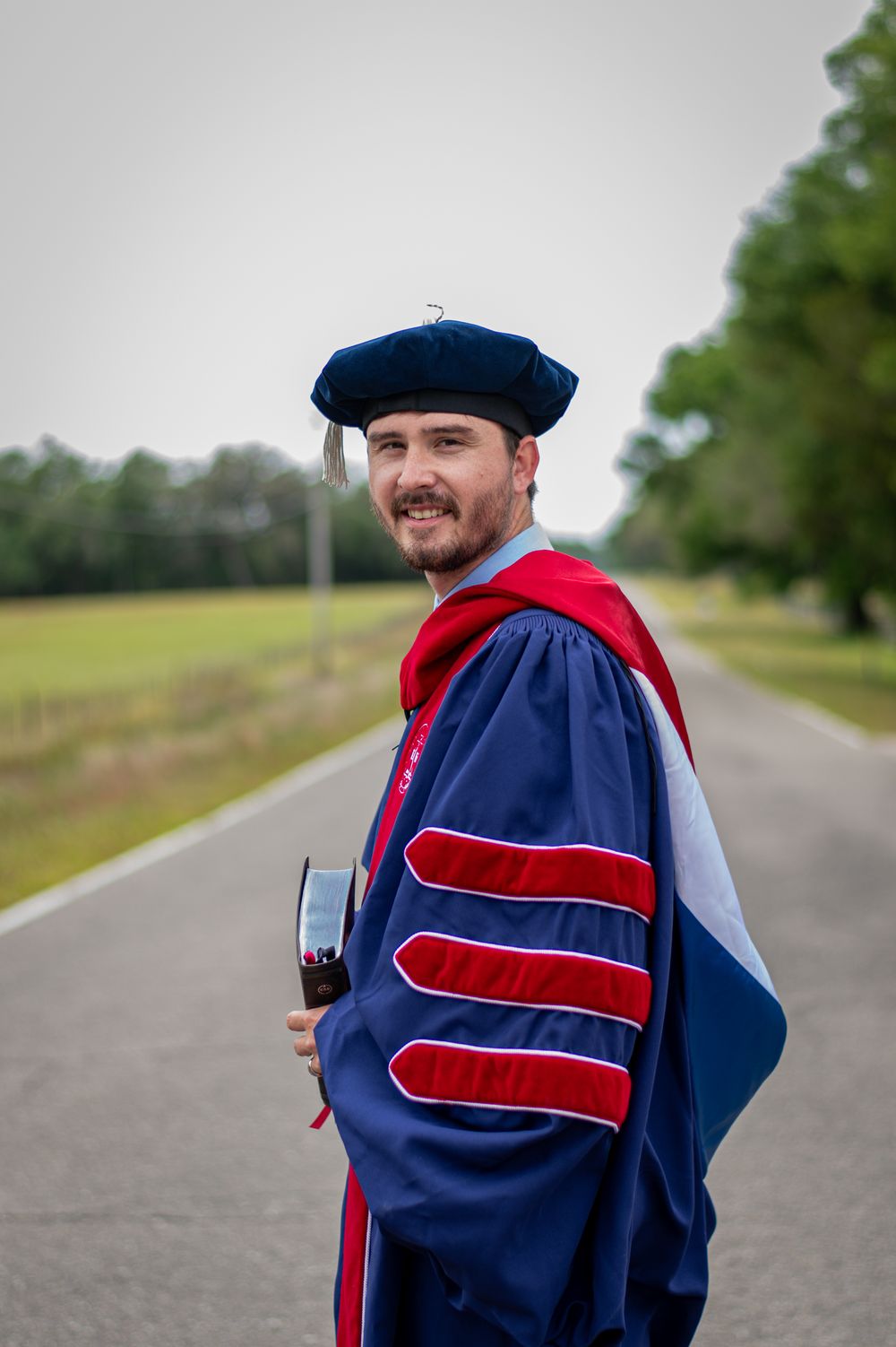 Graduate in blue and red doctoral robes holding diploma on road.