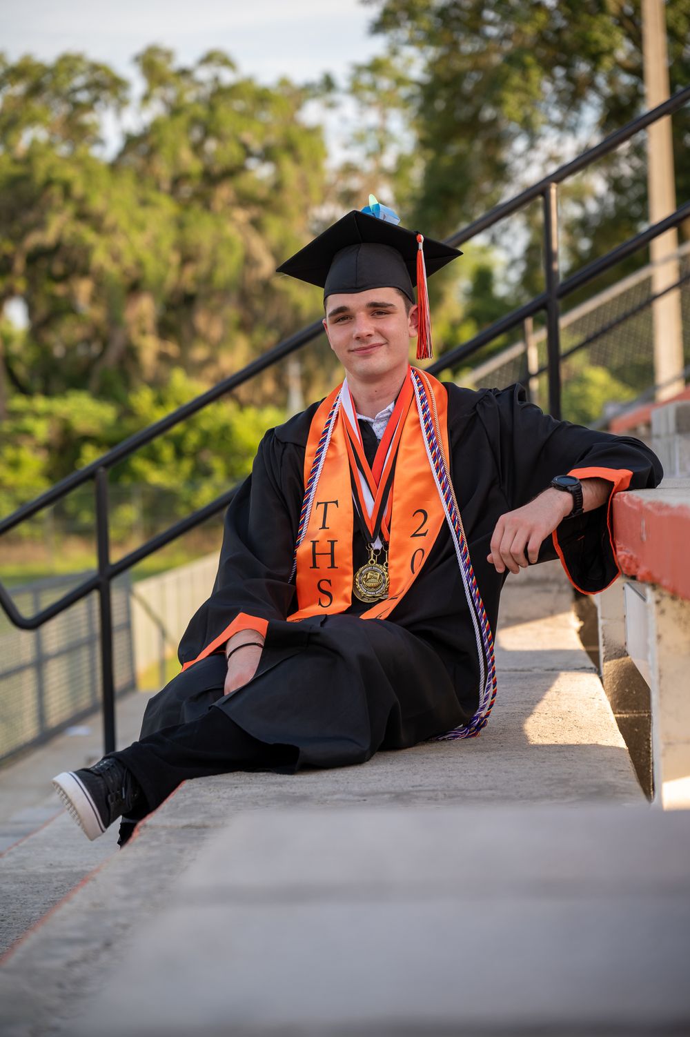 High school graduate in black cap and gown with orange THS stole and medal sits on bleacher steps smiling.