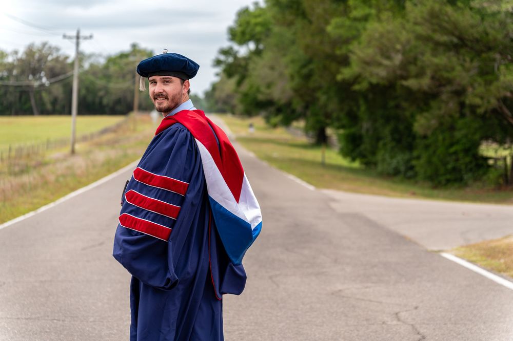Man in doctoral graduation gown and cap standing on a road, wearing navy blue and red academic regalia.