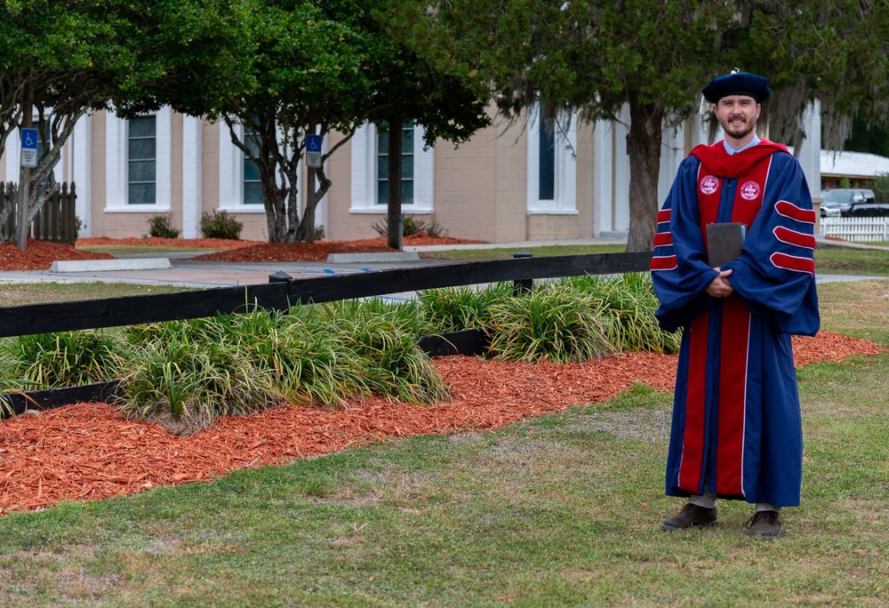 Man in doctoral graduation regalia stands proudly outdoors on a university campus on a cloudy day.