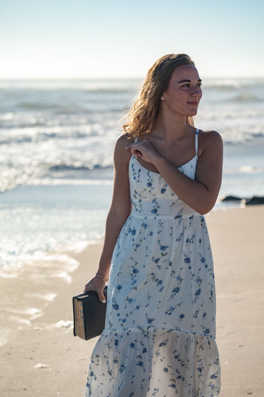 Woman in floral sundress holding a book on a sunny beach with ocean waves in the background.