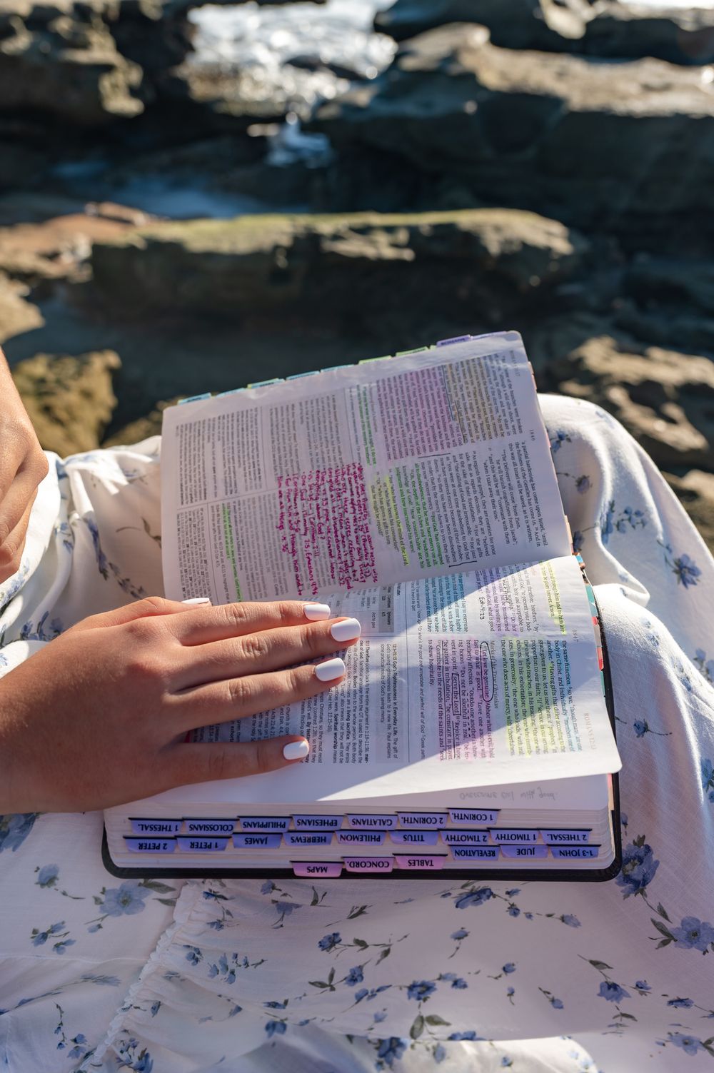 Woman reading a highlighted and annotated Bible outdoors near rocky shoreline, wearing a floral dress.