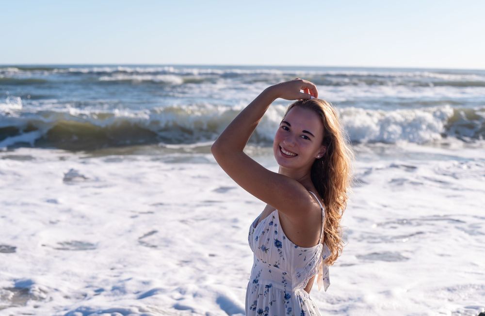 A senior in a floral dress walks along the beach shoreline with waves crashing behind her in summer sunset light.