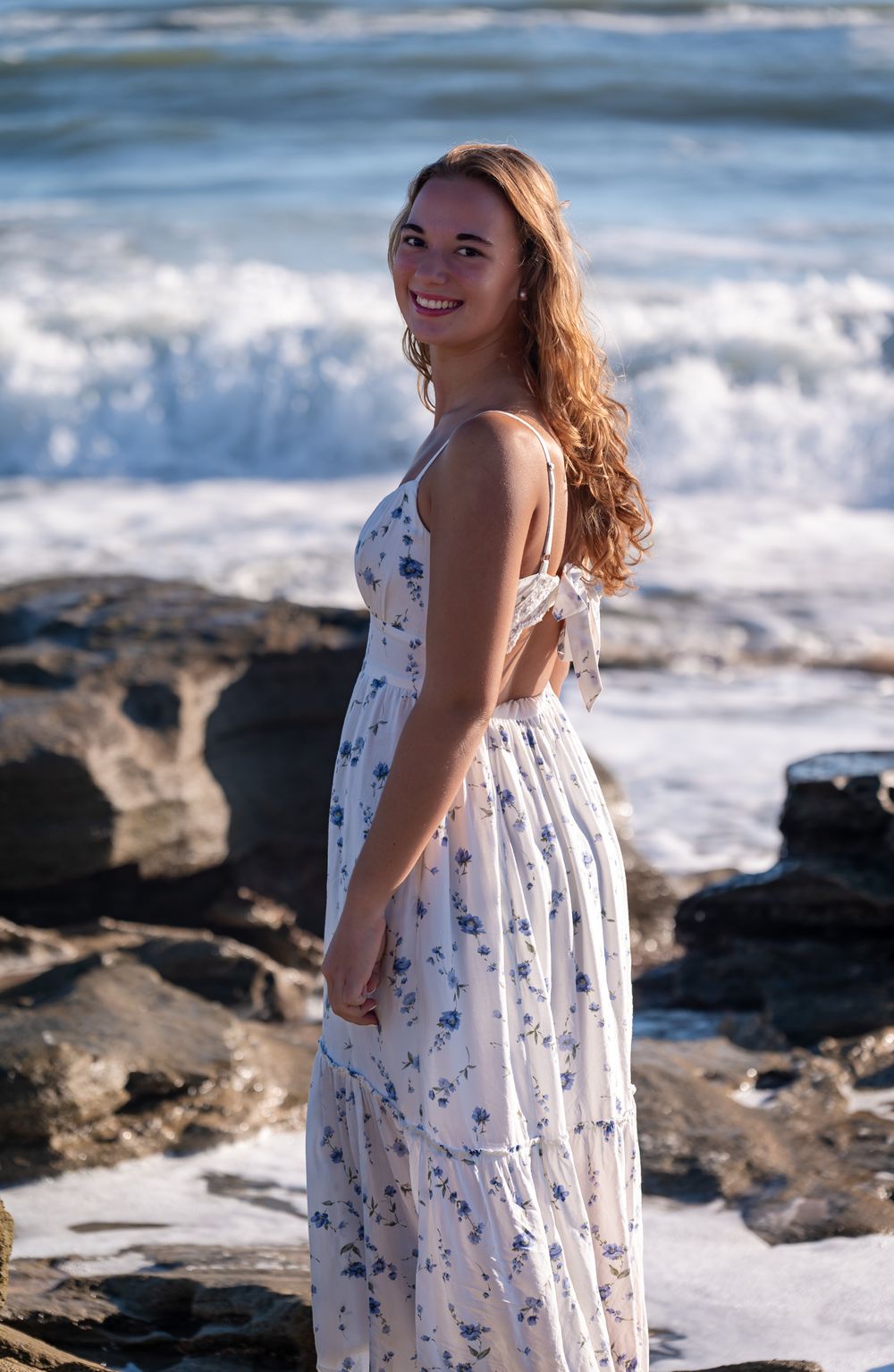 A senior in a flowy white floral sundress stands on rocky coastal shores with ocean waves crashing in the background.