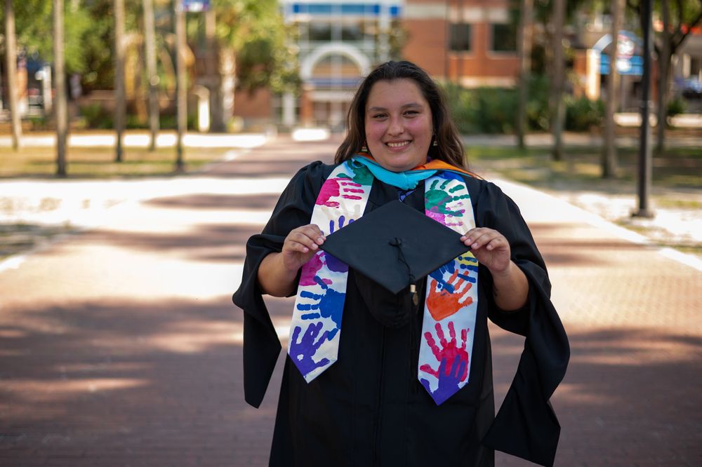 Graduate in cap and gown holds colorful handprint-decorated stole outdoors on a brick campus walkway.
