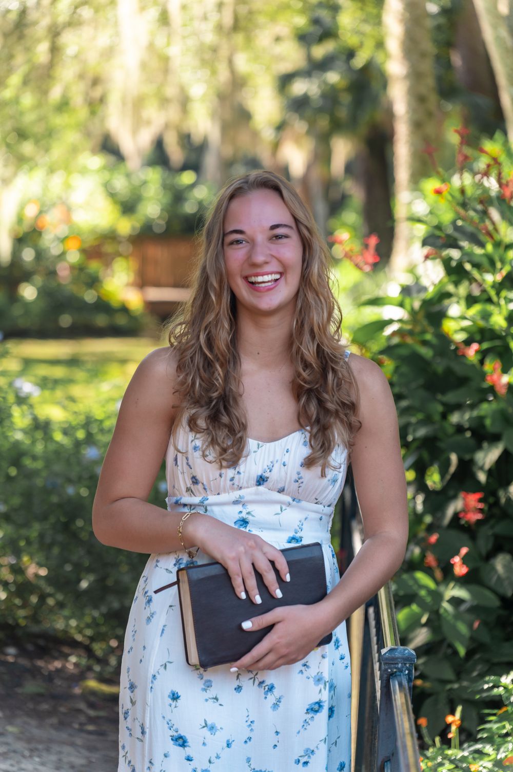 A senior in a white floral dress sits in a garden, holding a book and smiling warmly.