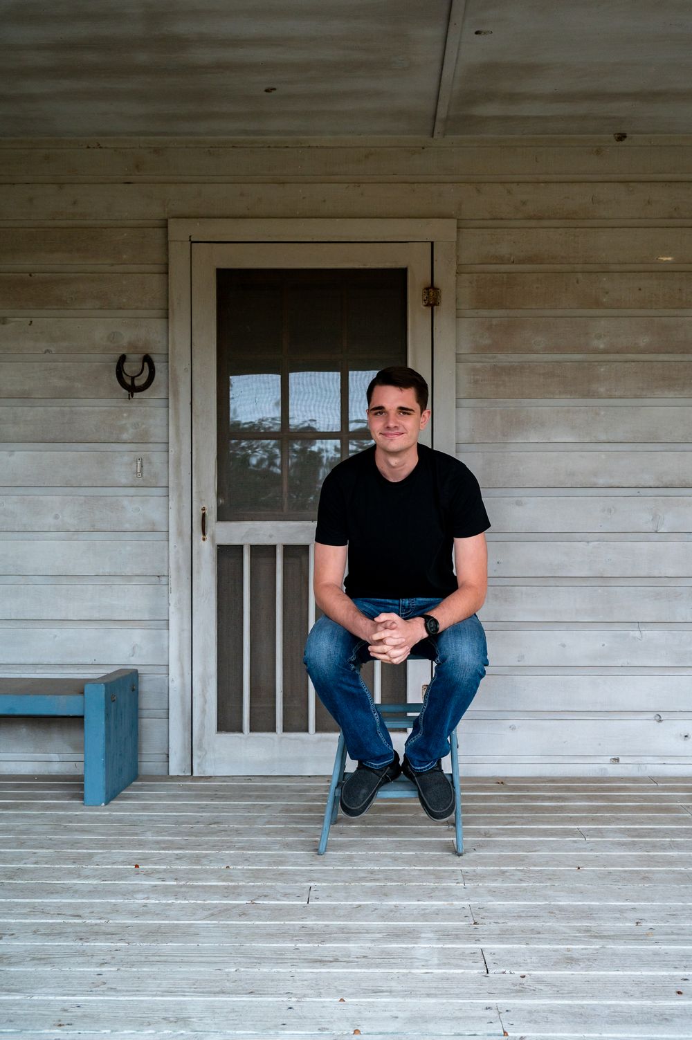 Young man in black t-shirt and jeans sitting on a rustic wooden porch chair outside a farmhouse door.