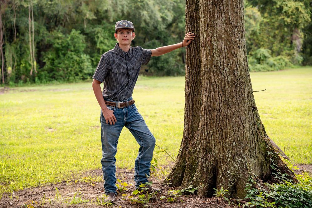 A person in casual western attire leans against a large tree trunk in a grassy outdoor setting.